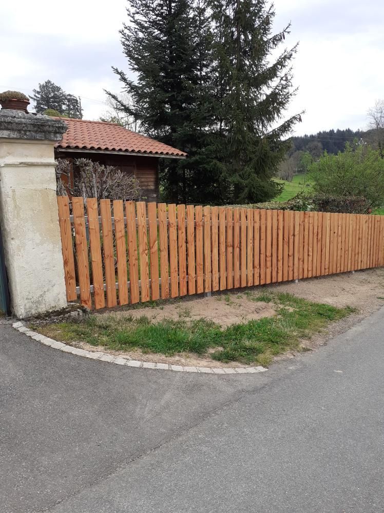 Clôture en bois le long d’une route, près d’une maison, avec un arbre et de la verdure en arrière-plan.
