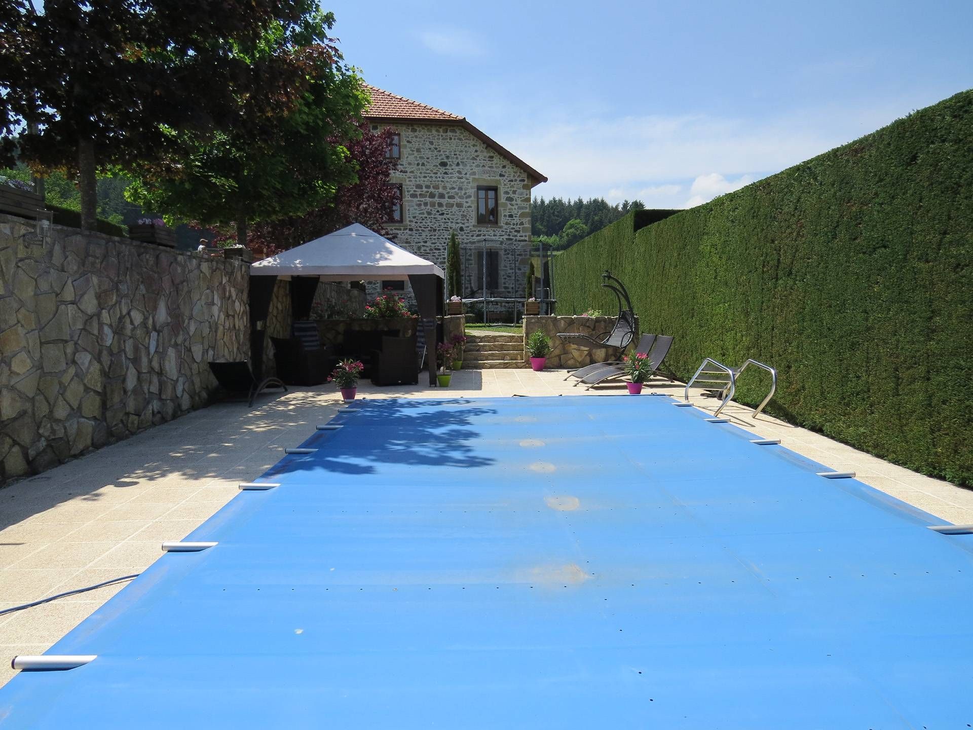 Piscine de jardin recouverte d’une bâche bleue, à côté d’une maison en pierre et d’une grande haie