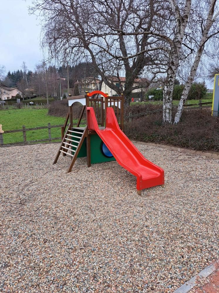 Toboggan rouge et structure d’escalade sur un sol en galets, dans un parc bordé d’arbres dénudés.