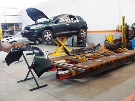 Un coche sobre un elevador en un taller mecánico, con el capó abierto y piezas del parachoques en el suelo.