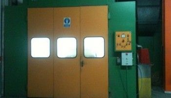Puertas dobles industriales de color naranja con pequeñas ventanas junto a una pared vegetal y un panel de control.