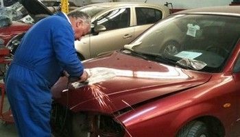 Un trabajador con mono azul repara la parte delantera de un coche rojo en un taller.