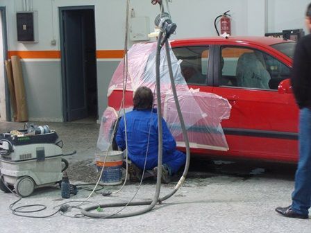 Dos personas pintan con aerosol un coche rojo en un taller, con mangueras y equipos cerca.