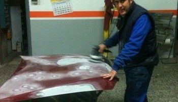 Un hombre lija un gran panel rojo en un taller; lleva una chaqueta azul y un chaleco negro.