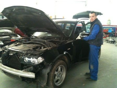 Mecánico con mono azul trabajando en un coche negro con el capó abierto en un garaje.