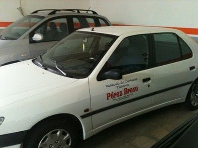 Un sedán blanco con el logotipo de “Pérez Bravo” estacionado en el interior, con un coche plateado detrás.