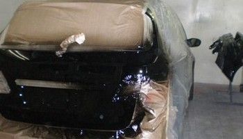 Coche negro envuelto en plástico transparente y acolchado color canela, aparcado en un garaje con otro vehículo cubierto cerca.