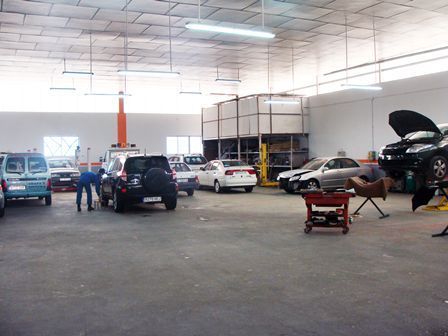 Taller mecánico con varios coches, un elevador y dos personas trabajando dentro de un amplio garaje.