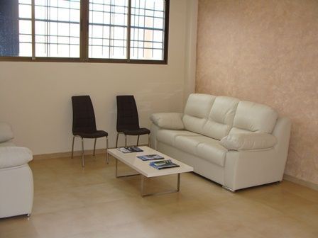 Sala de estar vacía con un sofá blanco, dos sillas negras y una mesa de centro baja junto a la ventana.