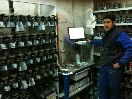 Un hombre está de pie junto a un ordenador en un almacén con estanterías llenas de artículos y equipos embalados.