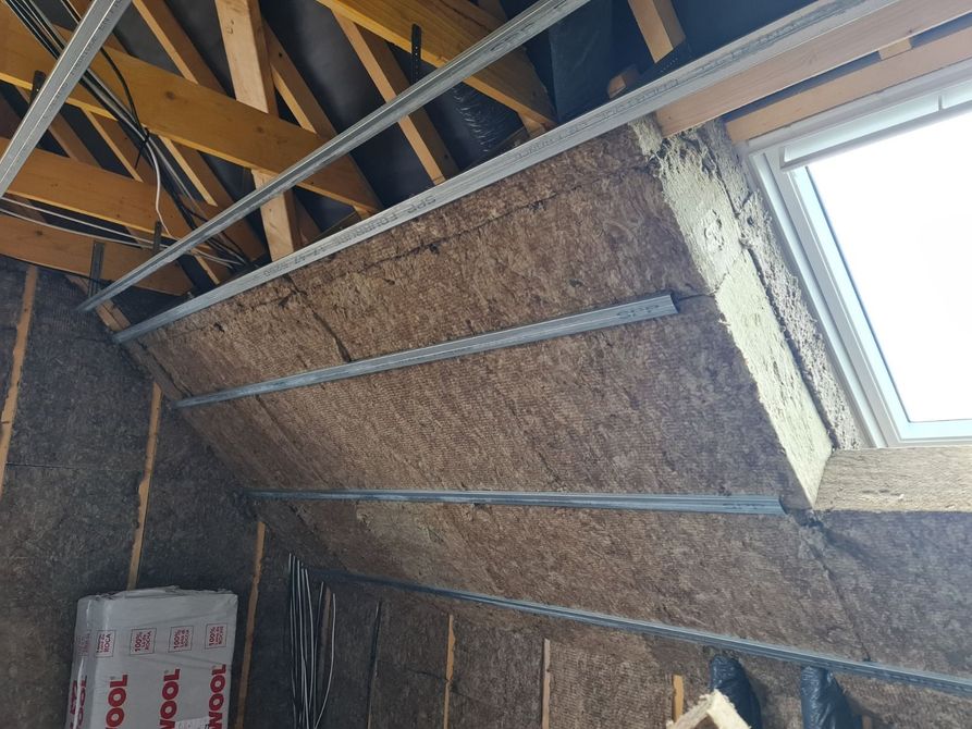 Mur intérieur en construction ; fenêtre au centre. L’isolation et les montants métalliques sont visibles. Une petite échelle est présente.