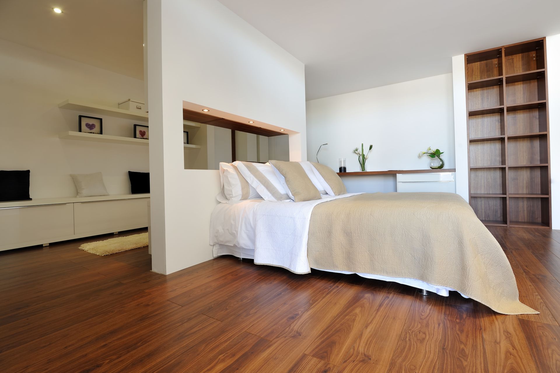 Chambre avec parquet, lit recouvert d'un couvre-lit beige, étagères et bureau intégrés, et murs blancs.