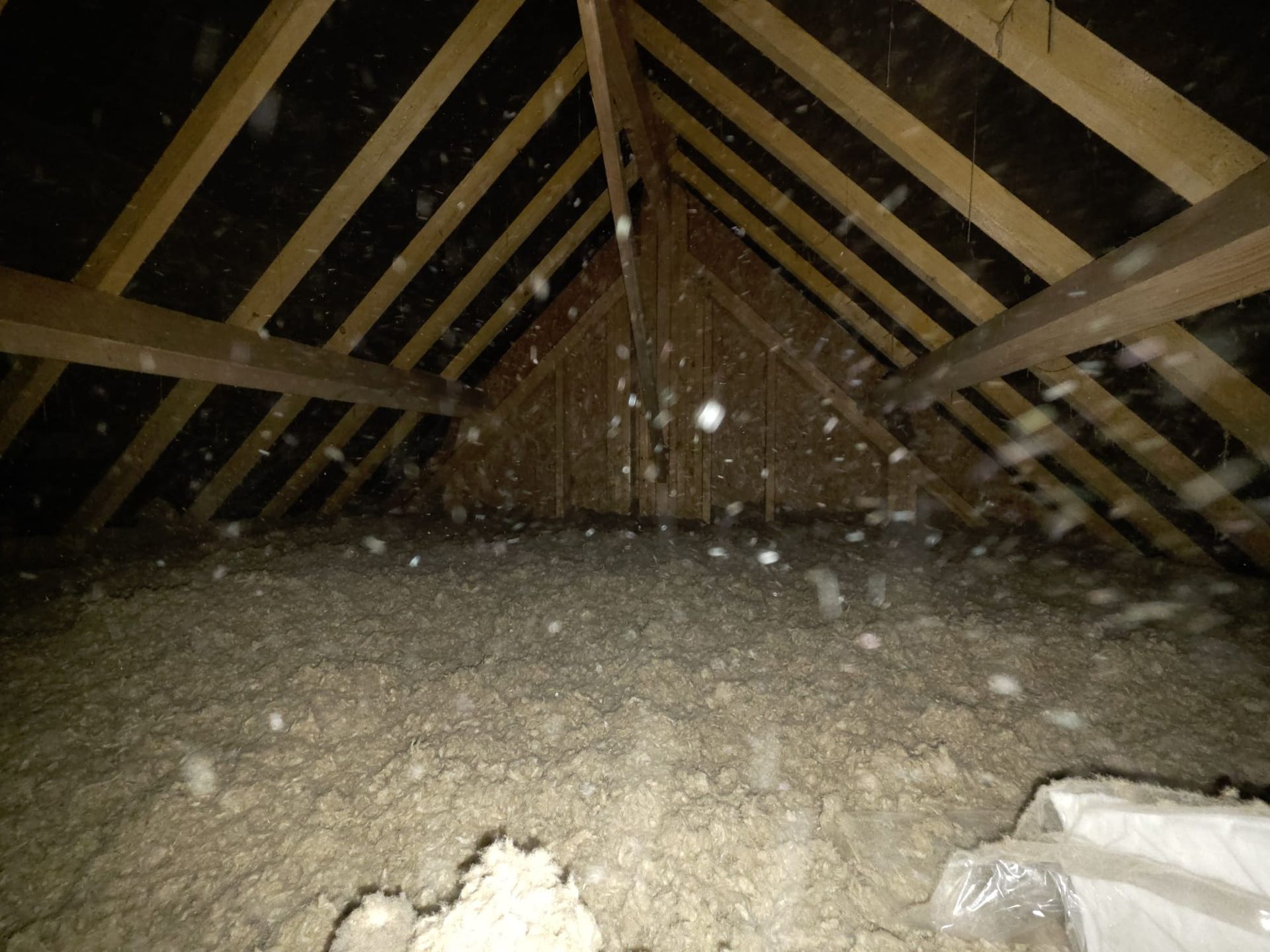 Intérieur du grenier avec poutres en bois et isolation. Des débris clairs et d'éventuelles particules de poussière sont visibles.