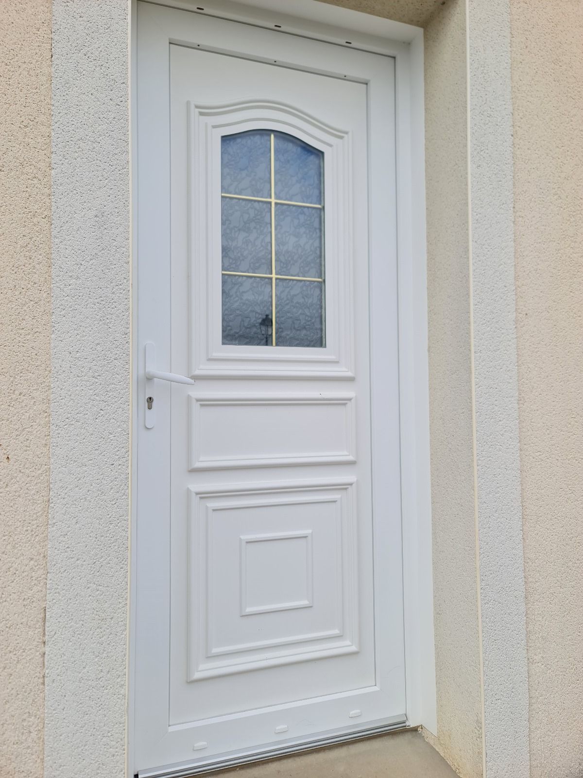 Porte d'entrée blanche avec fenêtre rectangulaire et panneaux décoratifs, encastrée dans un mur beige clair.