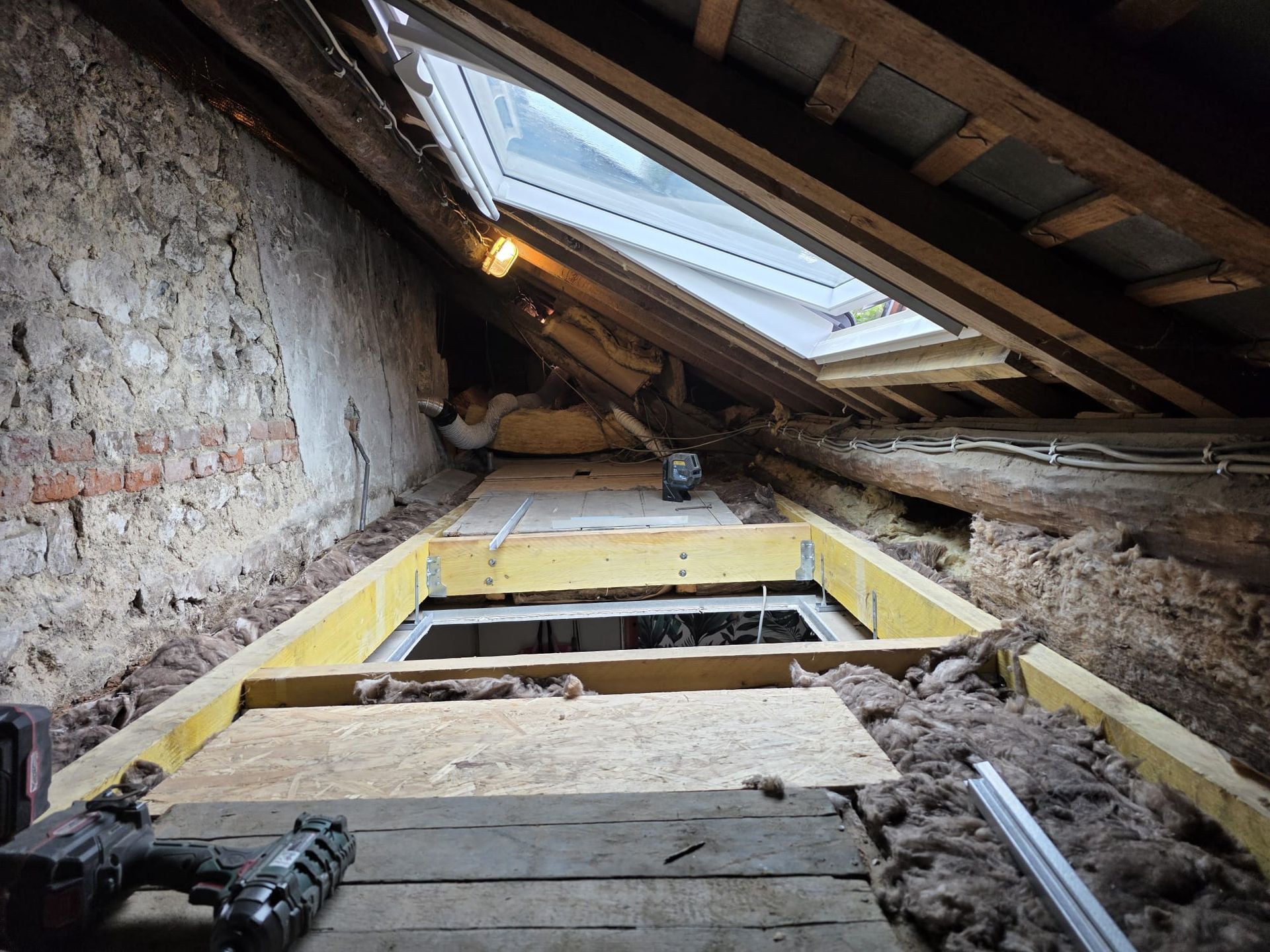 Intérieur du grenier avec puits de lumière ouvert, poutres en bois brut, isolation et trappe d'accès encadrée de bois jaune.