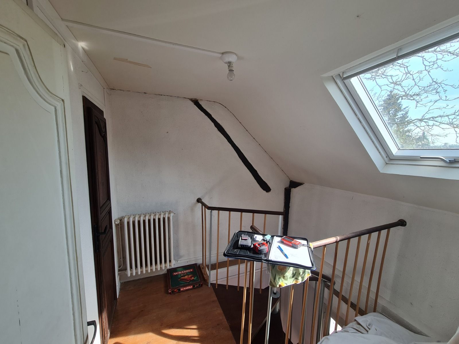 Pièce mansardée avec plafond mansardé, puits de lumière et rambarde. Porte et radiateur à gauche, bureau avec objets.