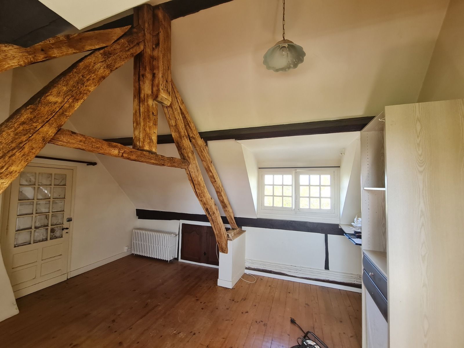 Pièce avec poutres apparentes, plafond mansardé, petite fenêtre et parquet.