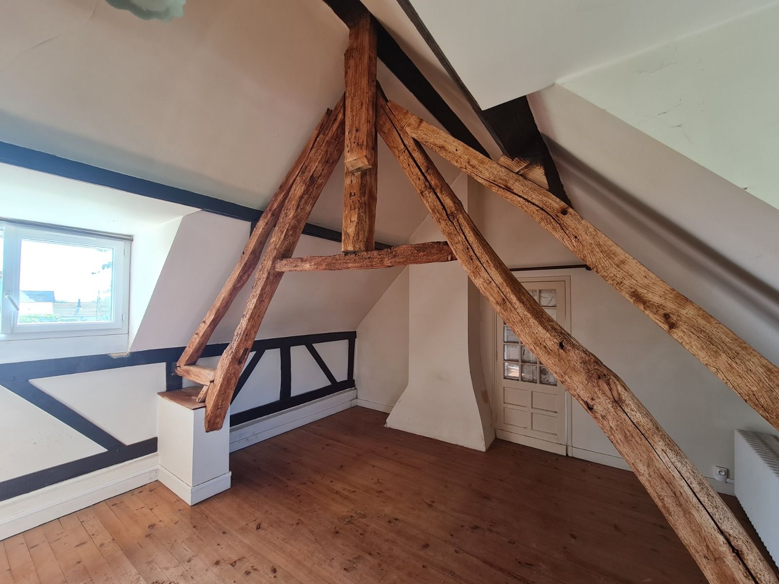 Pièce mansardée avec poutres apparentes, plafond mansardé, fenêtre et parquet.