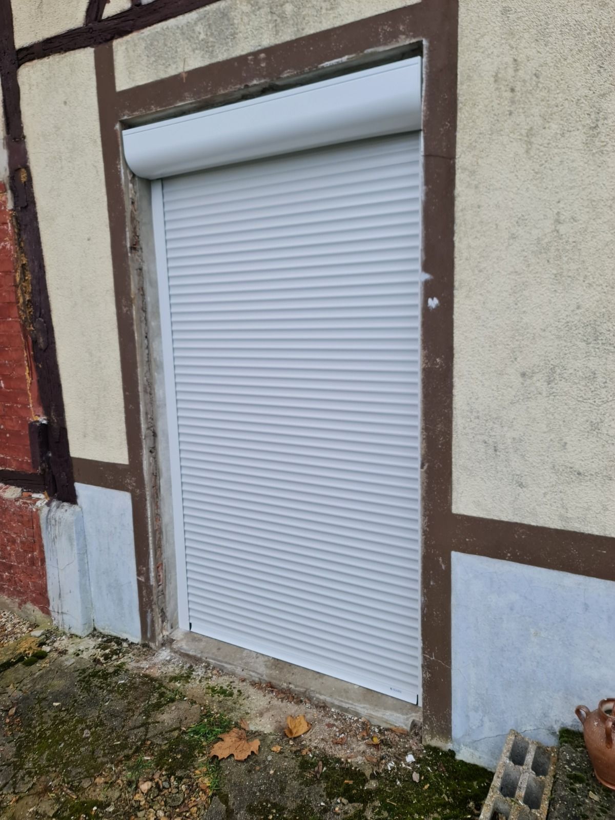 Vue extérieure d'un volet roulant gris clair fermé sur une porte encadrée par des matériaux de construction bruns et beiges.