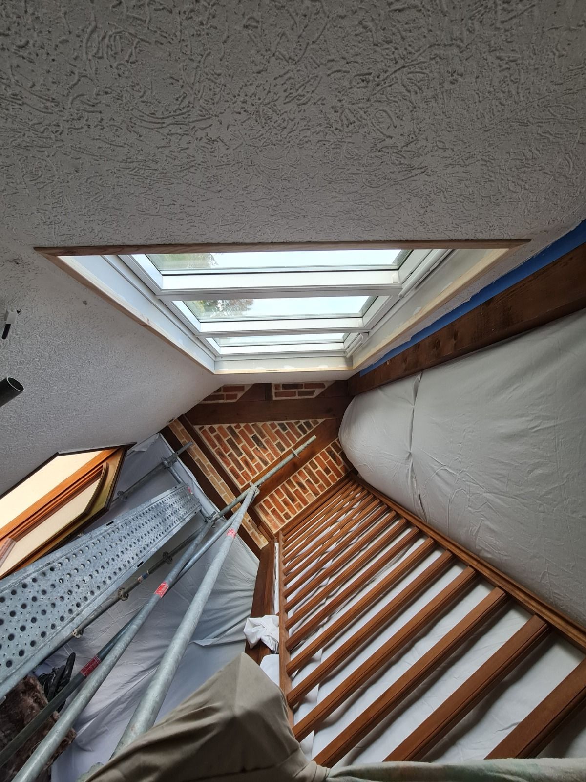 Vue plongeante sur une pièce avec puits de lumière, poutres apparentes et escalier. Tons blancs, beiges et bruns.