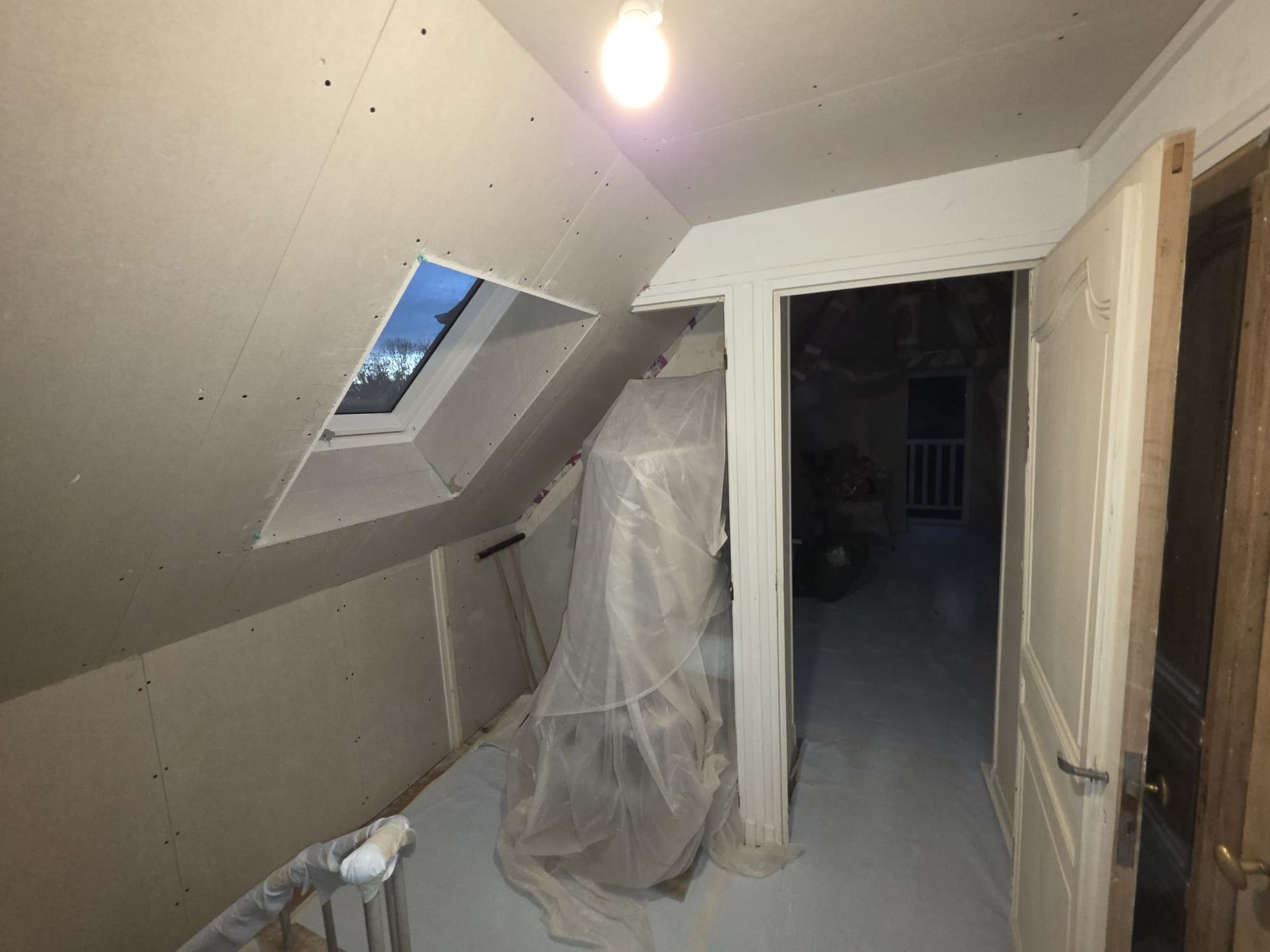 Pièce mansardée en cours d'aménagement avec murs inclinés, puits de lumière et porte donnant sur une pièce sombre.