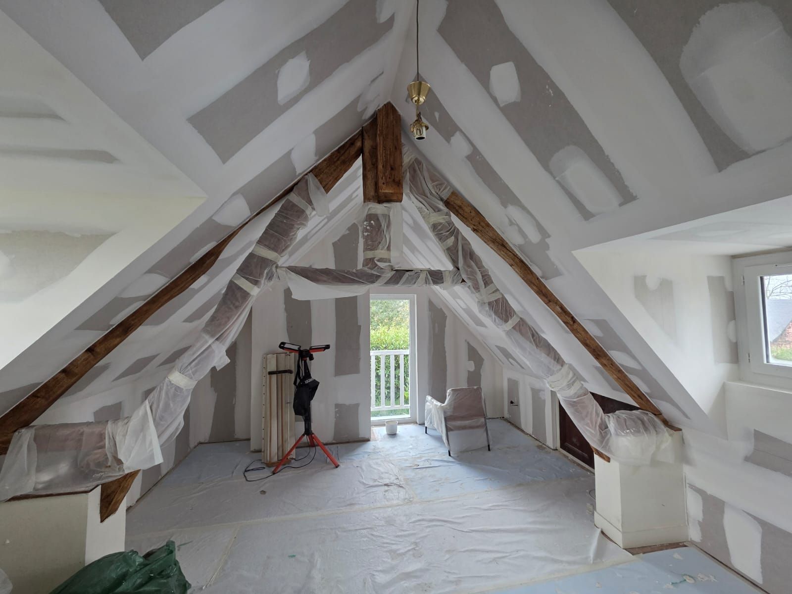 Aménagement intérieur des combles : poutres apparentes, plaques de plâtre et bâches en plastique. Une porte-fenêtre donnant sur un balcon est visible.
