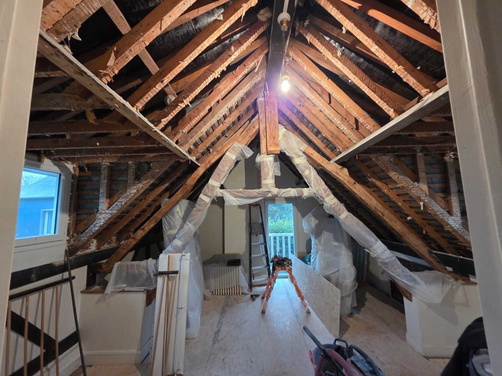 Vue intérieure d'un grenier non aménagé avec poutres en bois apparentes, murs blancs et matériaux de construction.