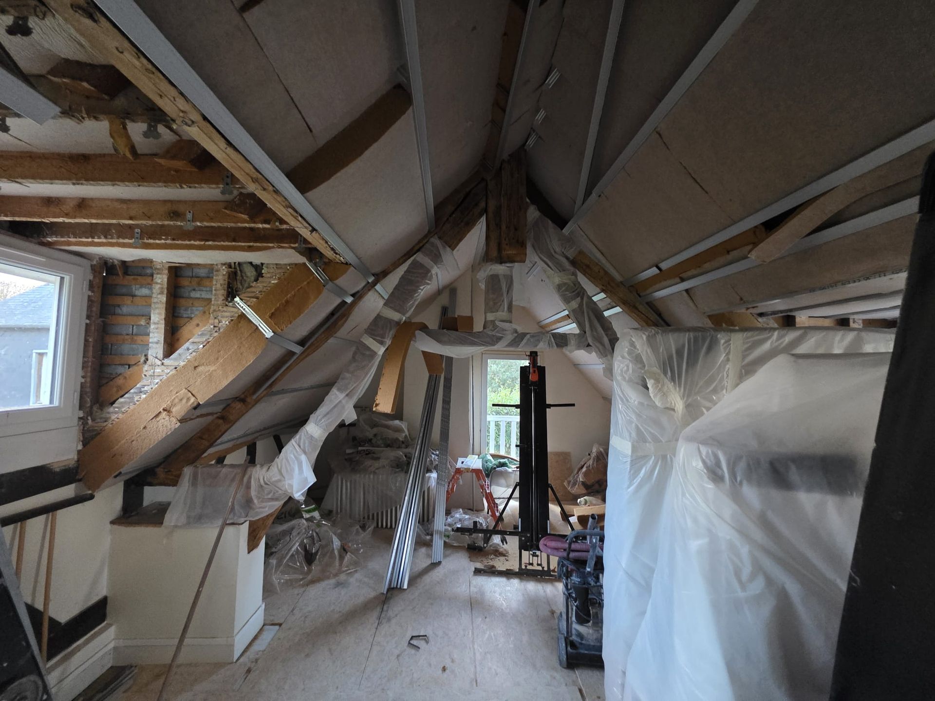 Intérieur d'un grenier en construction avec poutres apparentes, plaques de plâtre et bâches en plastique.