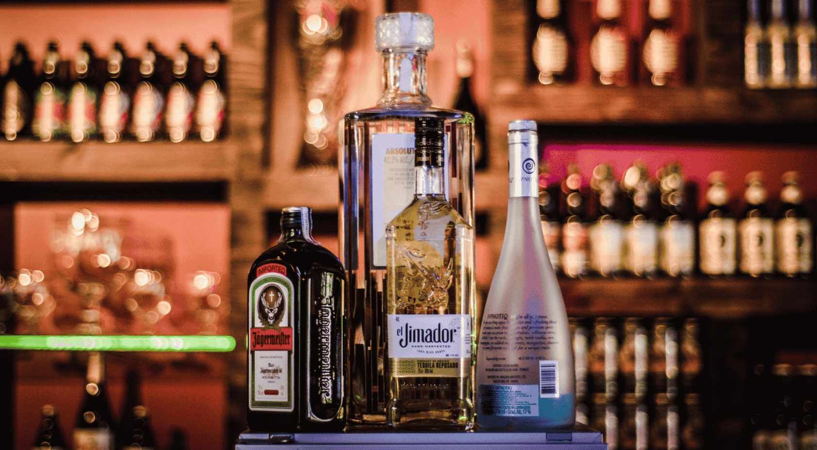Botellas de licor en un bar: Jägermeister, tequila y un aguardiente transparente. Estanterías con botellas al fondo.