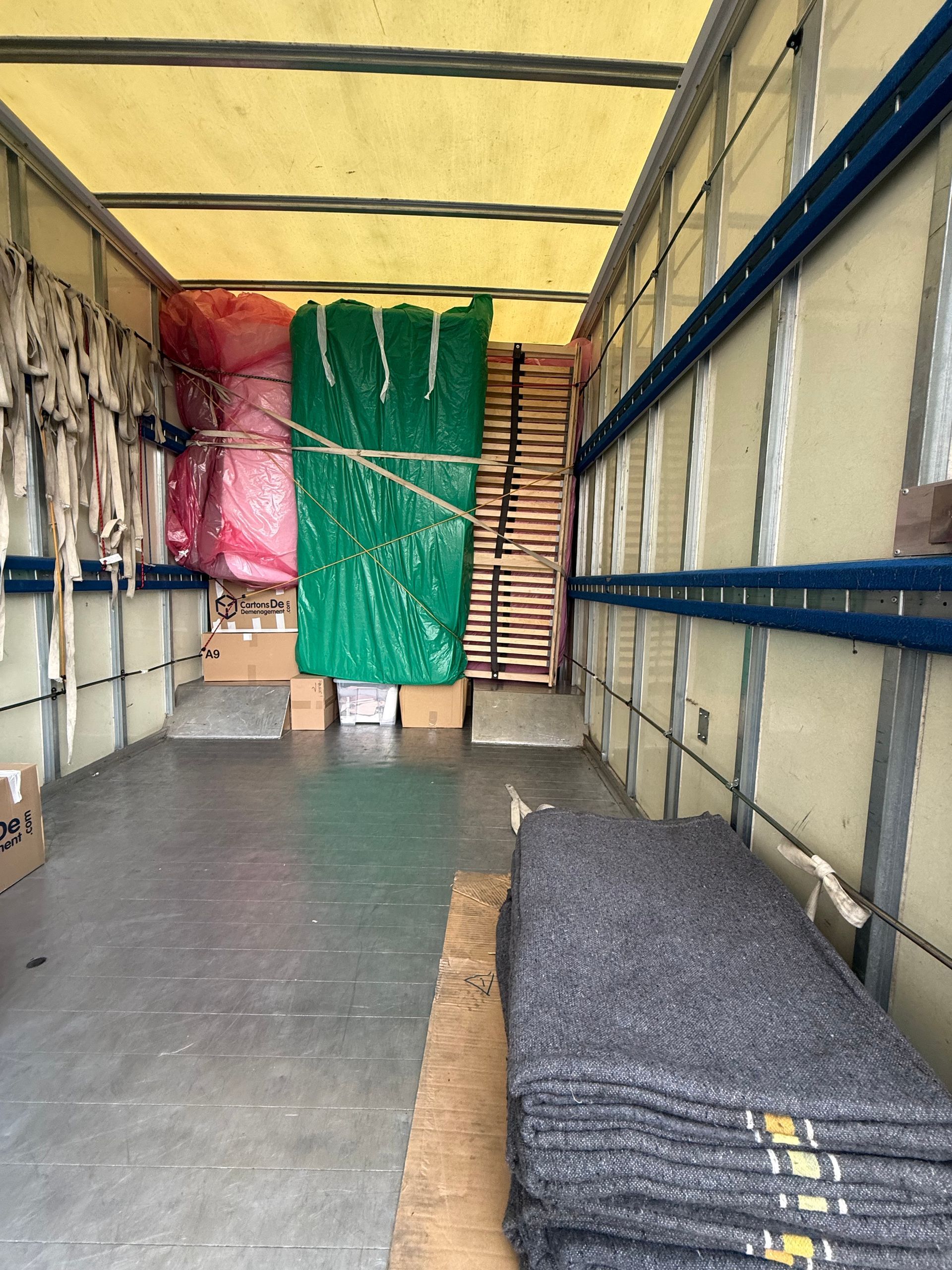 Intérieur d'un camion rempli de marchandises : des paquets emballés dans du plastique, des boîtes en carton et des couvertures grises