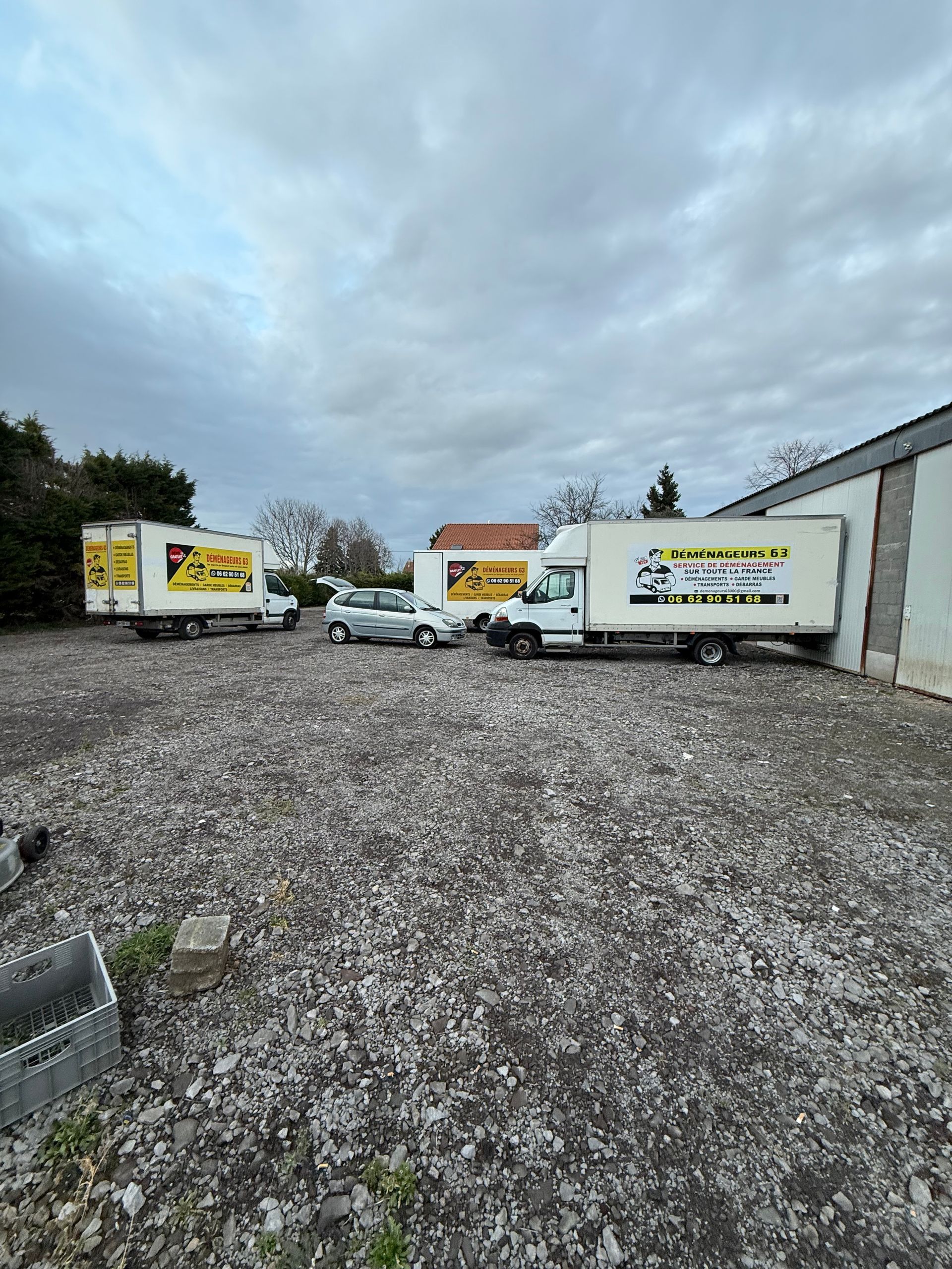 Trois camions de déménagement blancs stationnés sur un terrain gravillonné sous un ciel nuageux