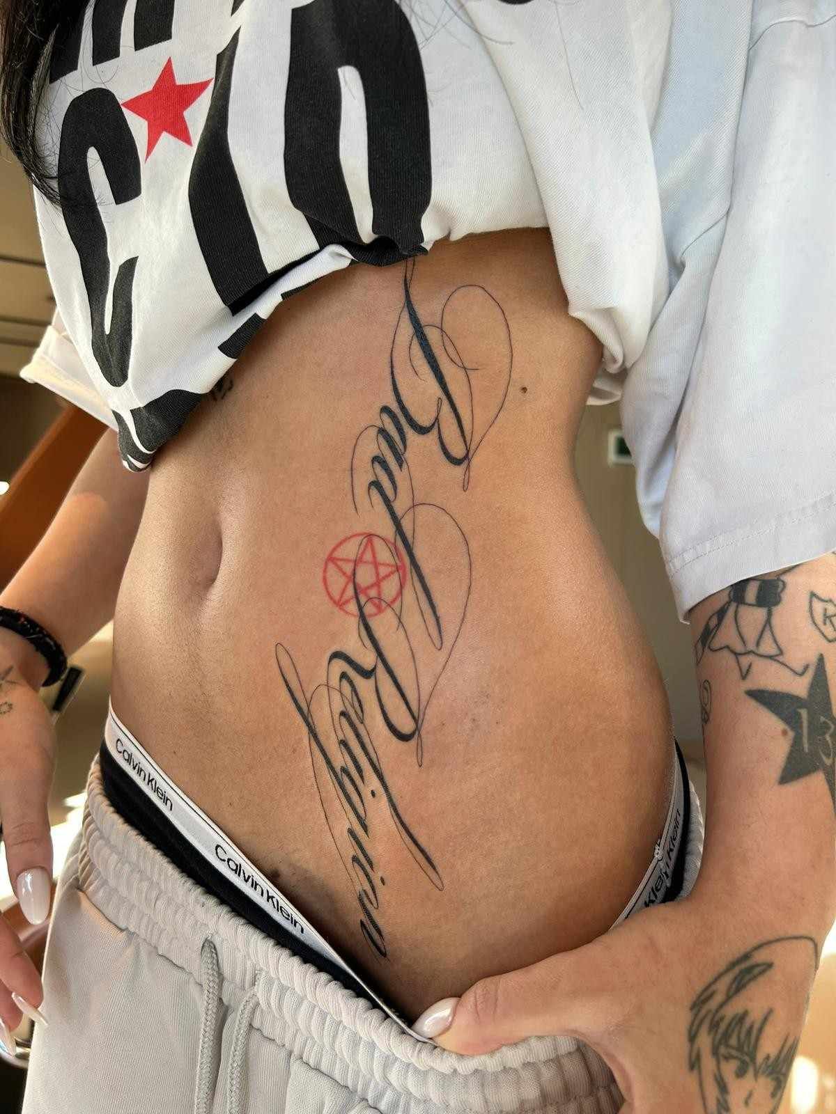 Persona con un tatuaje en el estómago, vestida con una camiseta corta y pantalones de tiro bajo, exponiendo parcialmente el tatuaje.