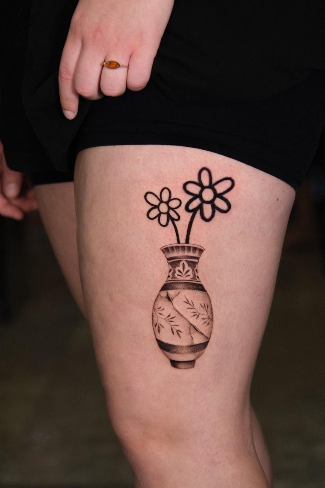 Tatuaje de un jarrón con flores en el muslo. Tinta marrón oscura.