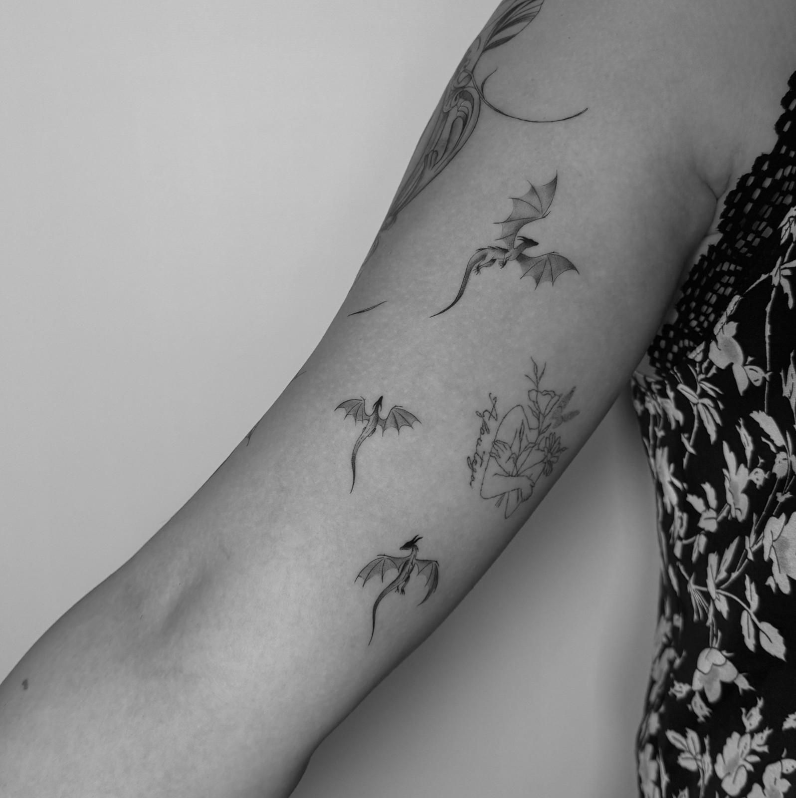 Brazo con tres pequeños tatuajes de dragón y un diseño floral; blanco y negro.