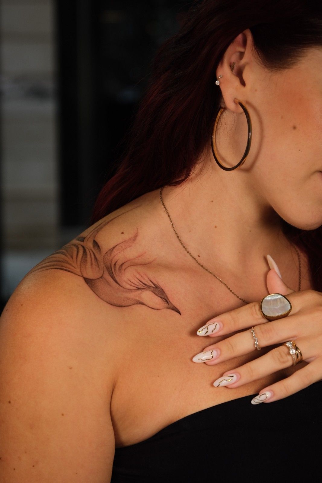 Mujer con tatuaje en el hombro, aros y anillos, vistiendo un top negro sin tirantes, posando en el interior.