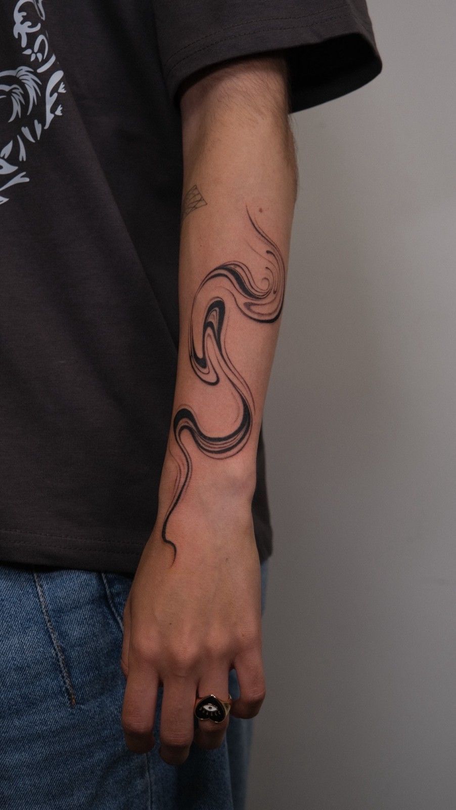 Brazo con un tatuaje de serpiente negra con pinceladas. La persona lleva vaqueros, camiseta oscura y un anillo.