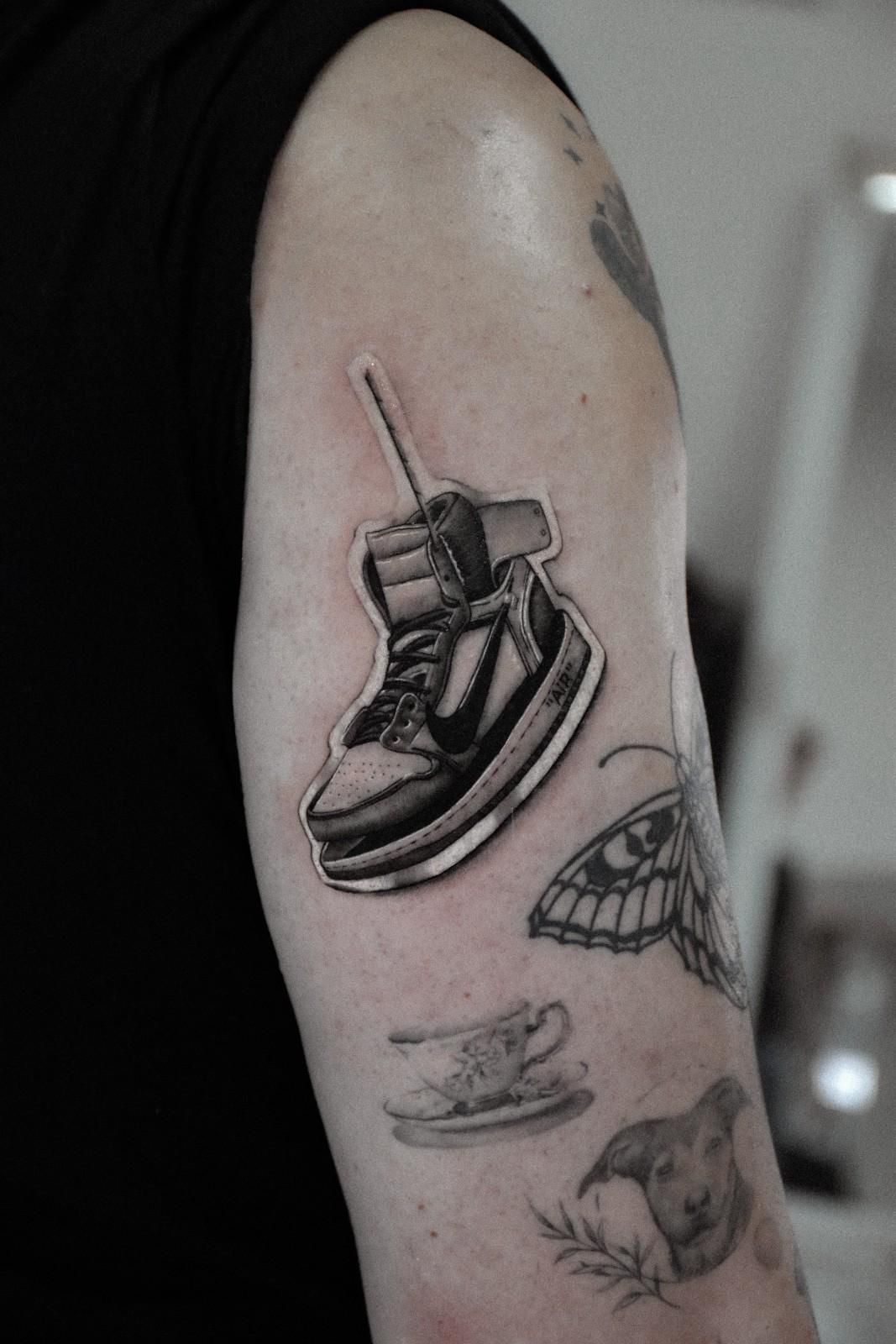 Brazo con tatuajes, entre ellos una zapatilla en escala de grises con un cordón, una polilla, una taza de té y un perro.