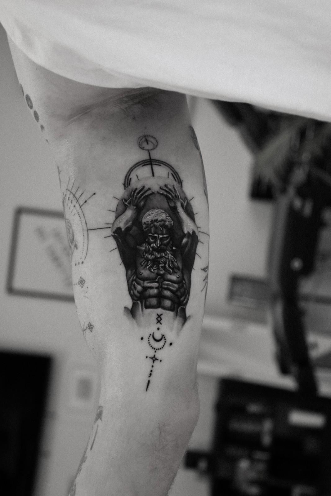 Tatuaje de una figura que cubre su rostro, rodeada de símbolos celestiales, en el brazo de una persona.