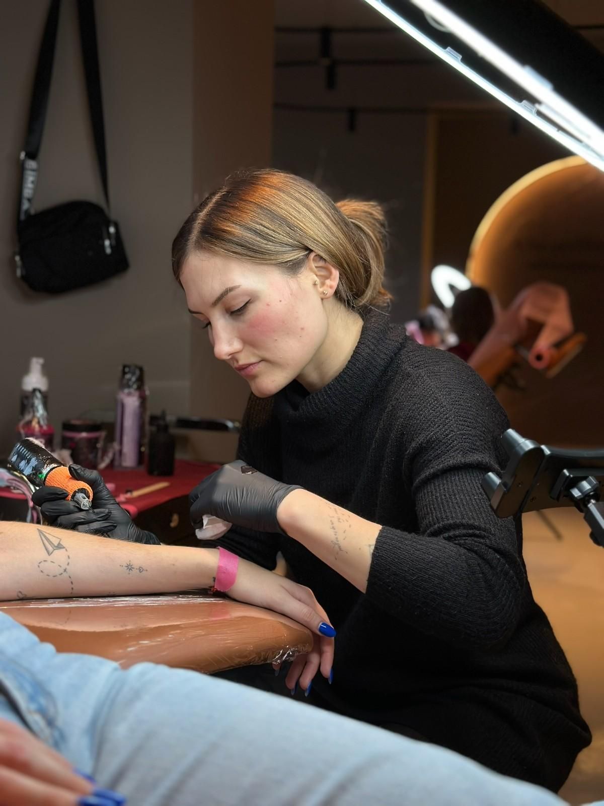 Tatuador con guantes negros, trabajando en el brazo de un cliente en una mesa. Iluminación brillante.