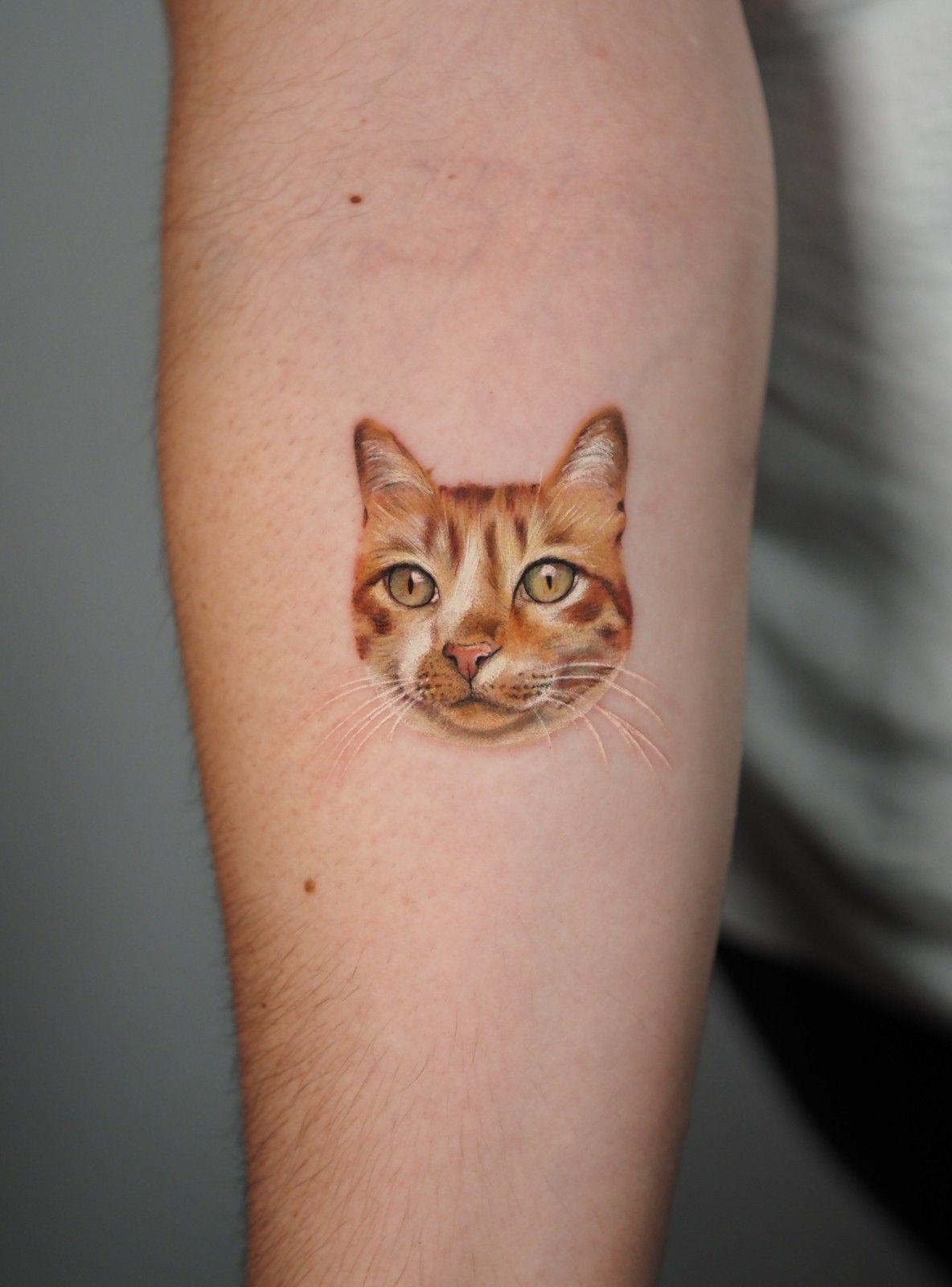 Tatuaje de la cara de un gato atigrado naranja en el brazo. El gato tiene ojos amarillos y bigotes visibles.