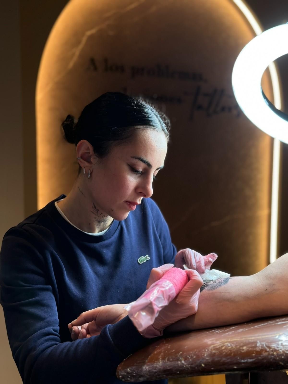 Artista del tatuaje con camisa azul y guantes, tatuando el brazo de un cliente con una pistola rosa. La luz de un anillo ilumina la escena.