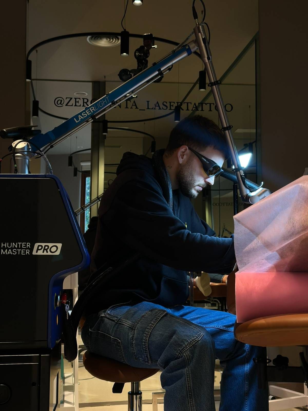 Hombre con gafas protectoras, trabajando en la eliminación de un tatuaje. Maquinaria azul y negra, tonos neutros.