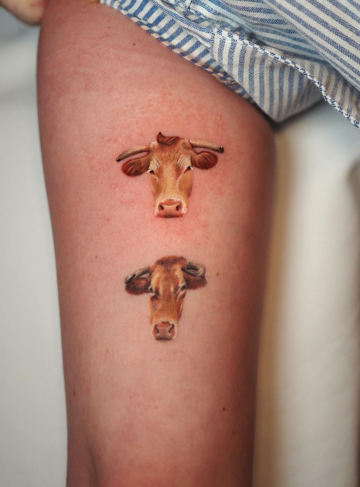 Dos tatuajes detallados de cabeza de vaca en una pierna, de color marrón claro y tostado.