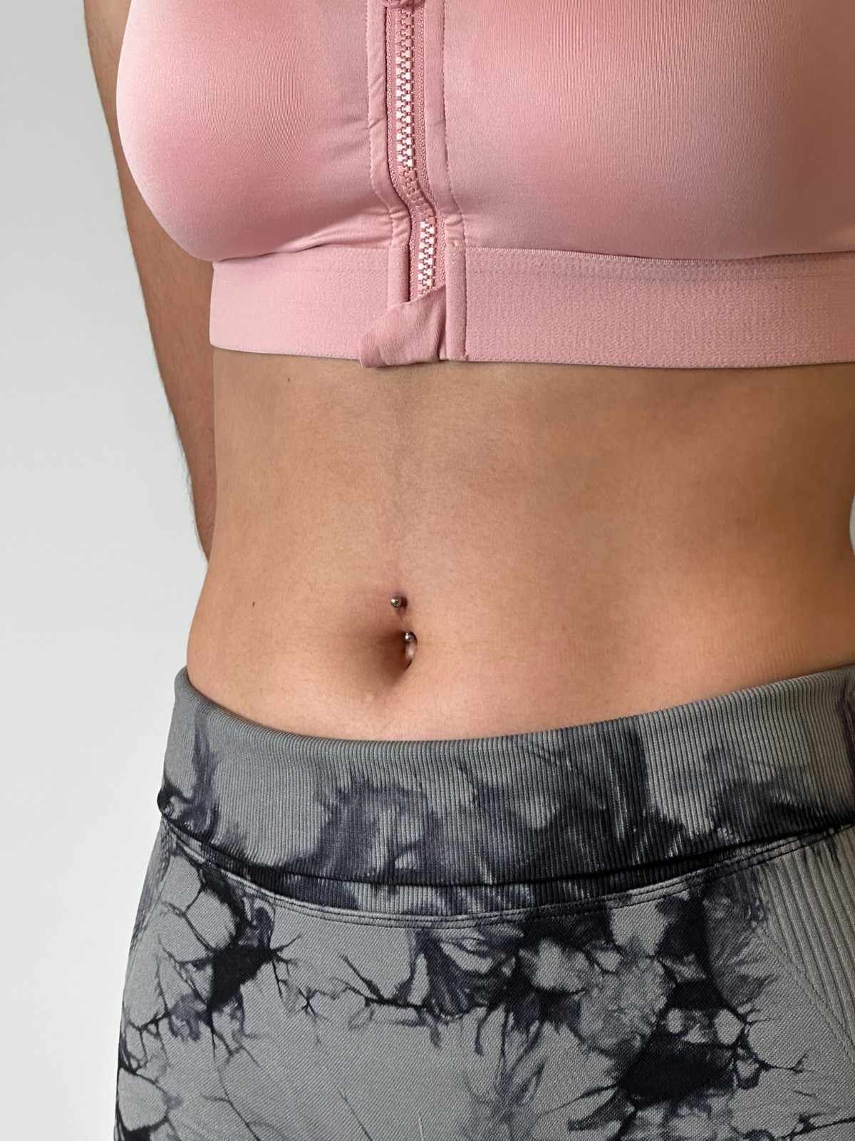 Torso de una persona que viste un sujetador deportivo rosa y mallas estampadas en gris y negro; piercing en el ombligo visible.