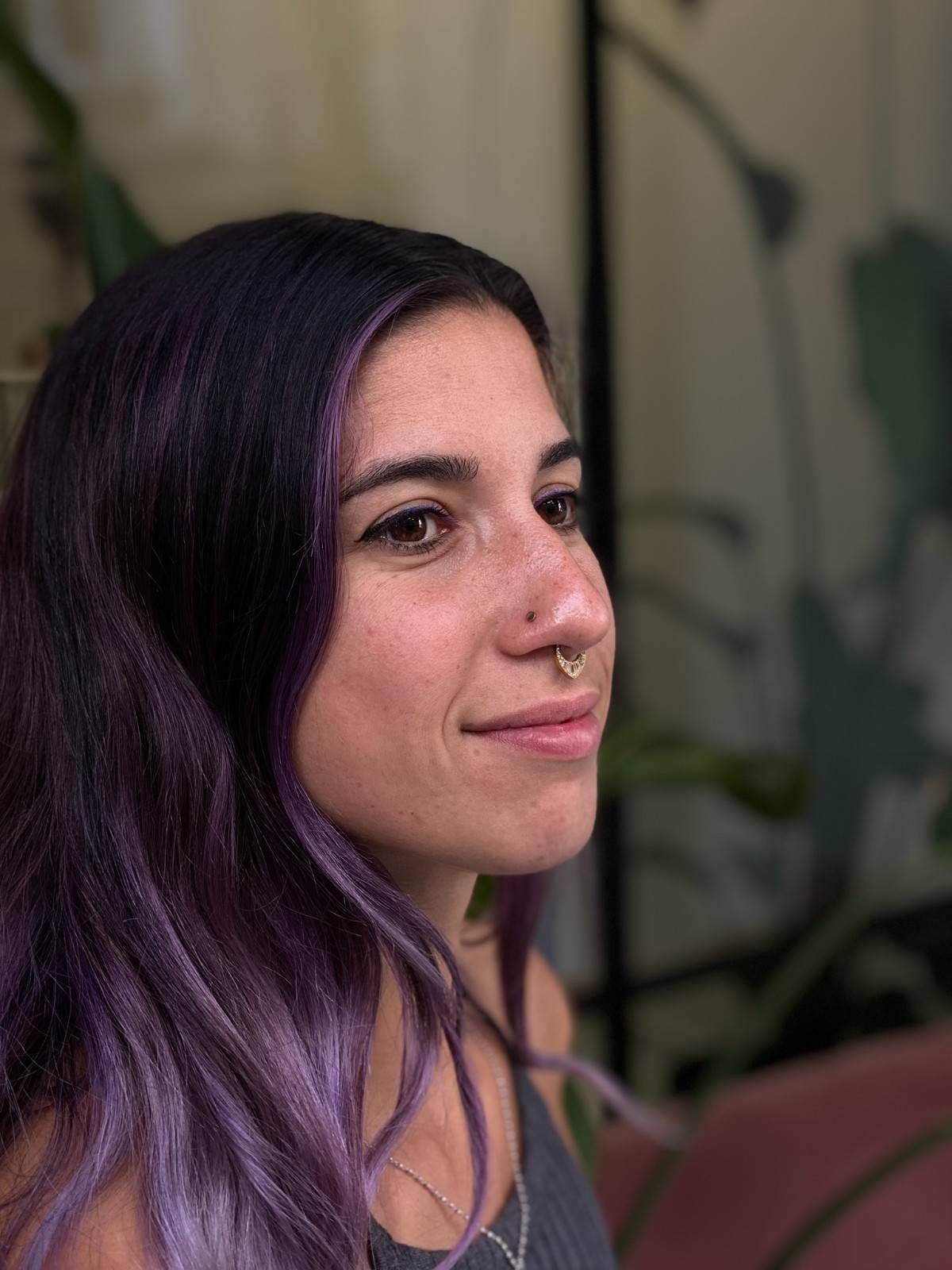 Mujer con cabello morado oscuro, piercing en el tabique, sonriendo, delante de una planta.