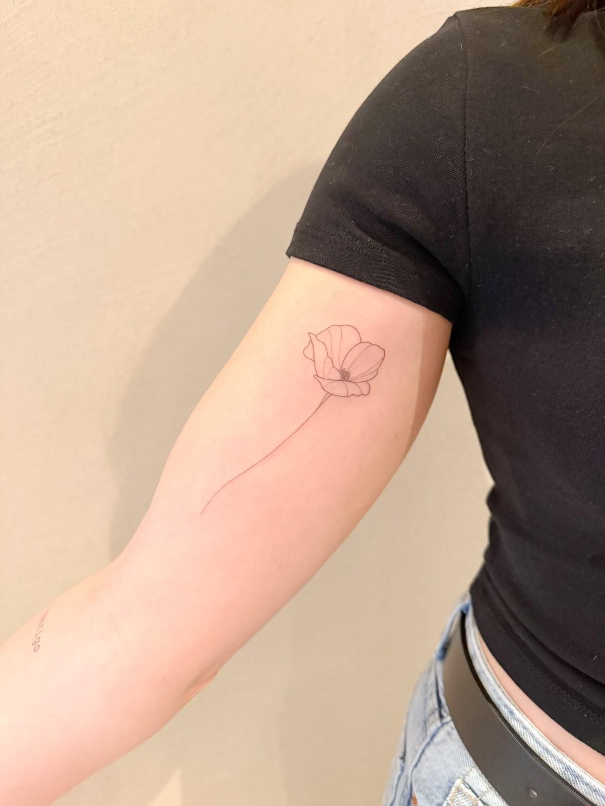 Brazo con un tatuaje minimalista de flor. La tinta negra delinea una amapola. La persona lleva una camisa negra y vaqueros.