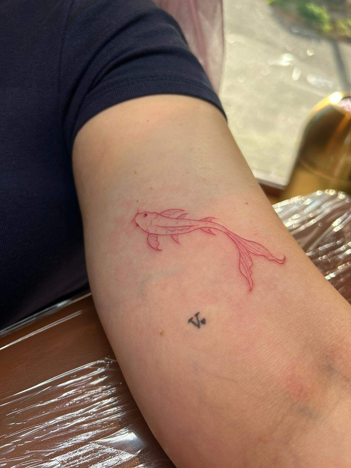 Tatuaje de un pez koi con contorno rojo en el antebrazo. Pequeño tatuaje negro debajo.