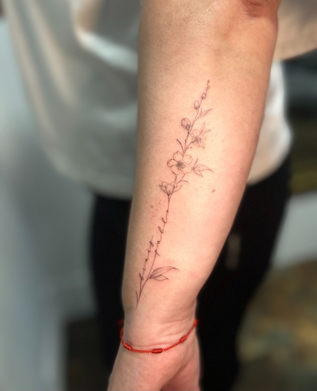 Tatuaje de flores delicadas y escritura en el antebrazo. Pulsera de hilo rojo en la muñeca.