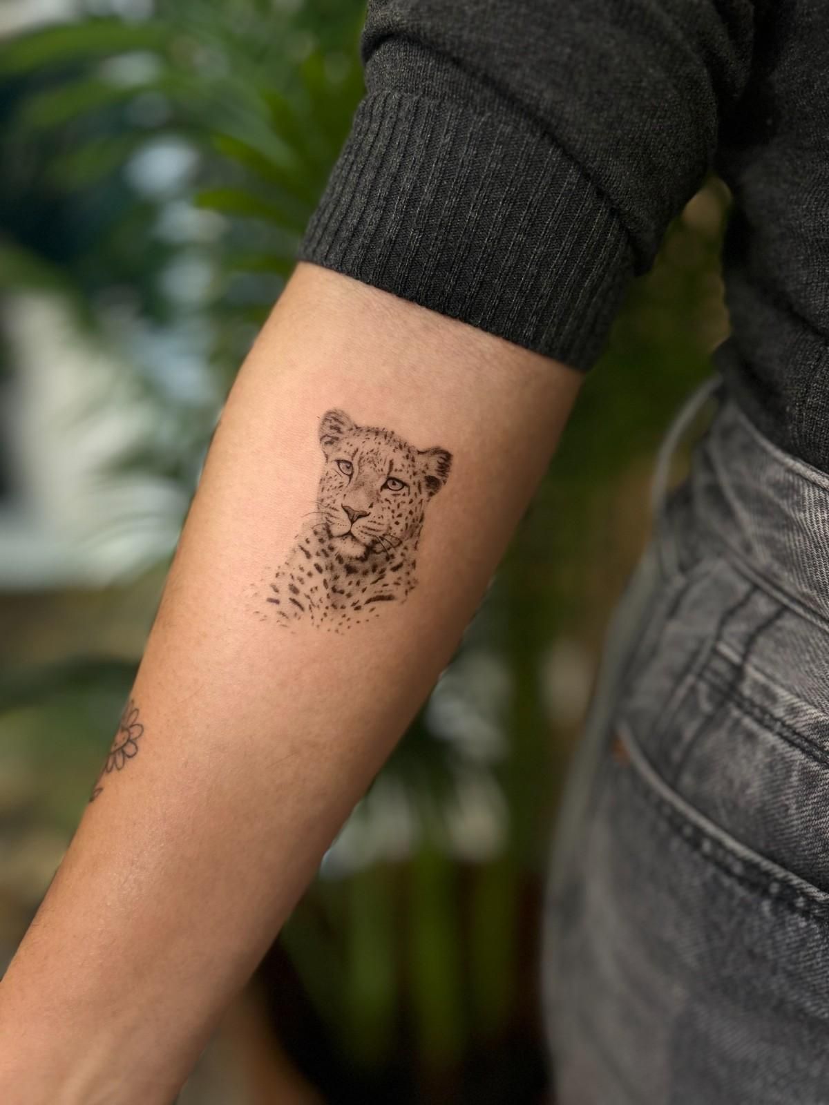 Tatuaje de una pequeña cara de león en el antebrazo de una persona que viste un suéter gris.