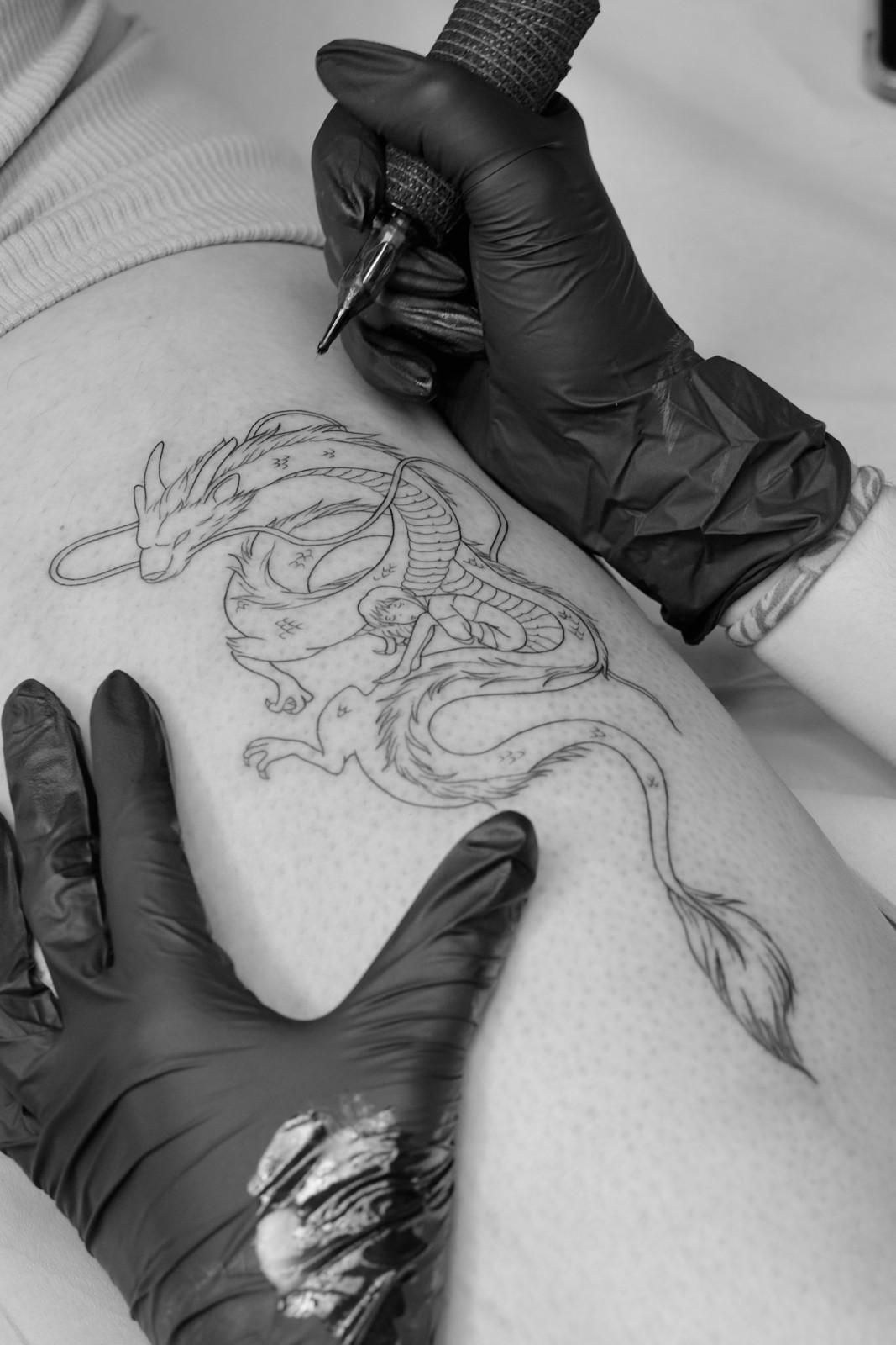 Artista del tatuaje con guantes negros, tatuando un diseño de dragón en la pierna de una persona.